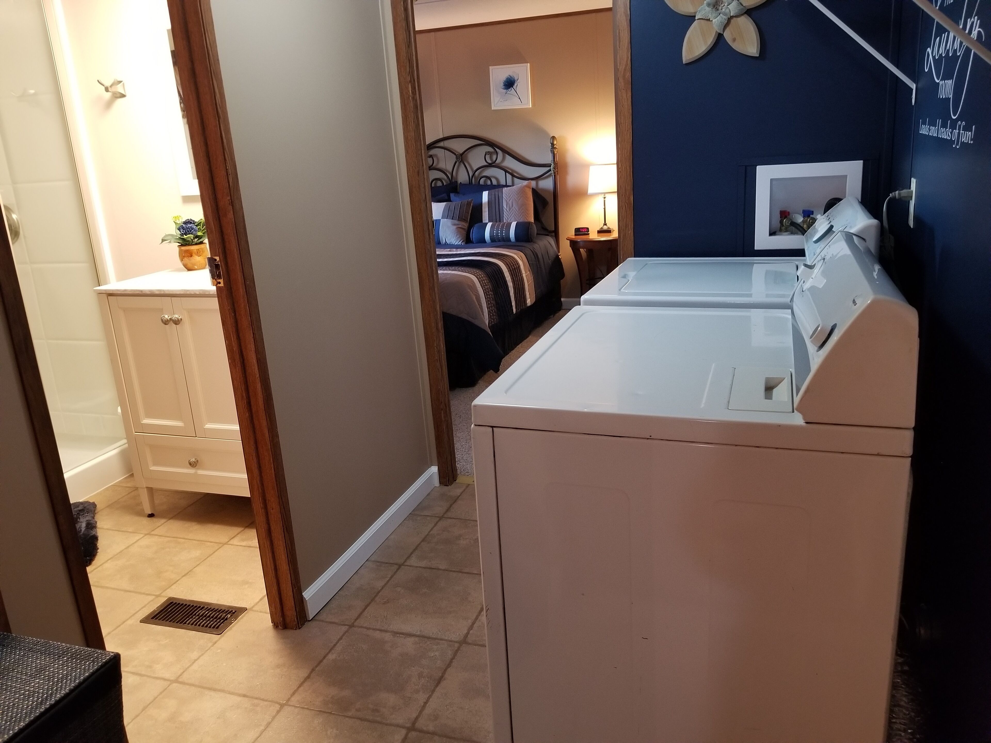 A laundry area with a washer and dryer, a bed with a headboard, a bedside table with a lamp, and a small plant on a cabinet.