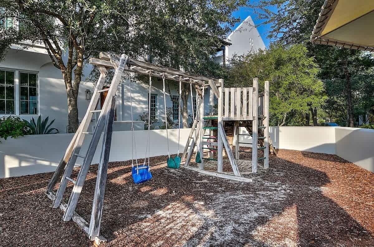 A playground with a swing set and climbing structure, surrounded by trees and mulch.