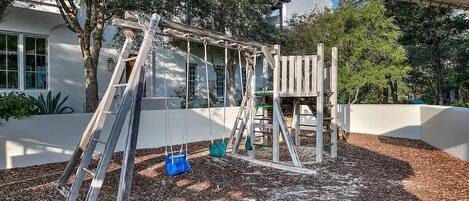 A playground with a swing set and climbing structure, surrounded by trees and mulch.