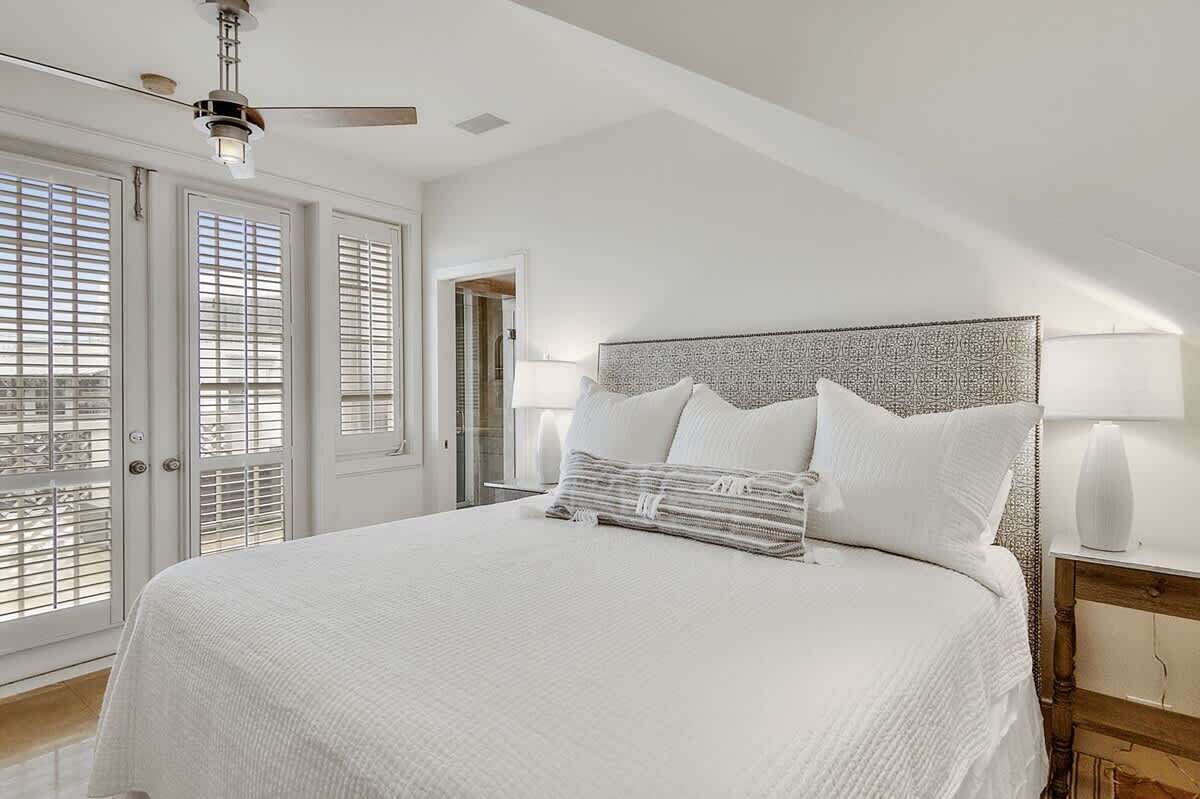 A bedroom with a large bed, a ceiling fan, and a view through the window blinds.