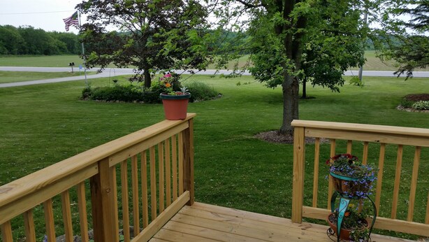 A wooden deck with a railing, a potted plant, and a view of a grassy area with a road and trees in the background. - Lime Ridge Guest House  (Elkhart Lake)