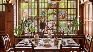 A dining area with a table set for four, surrounded by wooden chairs. There are potted plants on the windowsill, and a stained glass window depicting a scenic landscape. - Altes Gasthaus Kuhr (Papenburg)