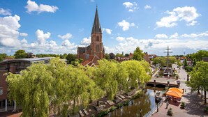 A historic church with a tall spire, a canal with a bridge, and a row of trees in the foreground. - Altes Gasthaus Kuhr (Papenburg)