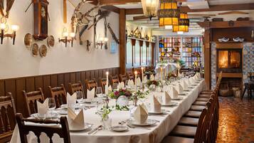 A long dining table set with white tablecloths, candles, and floral centerpieces in a warmly lit room with wooden beams and a fireplace.
