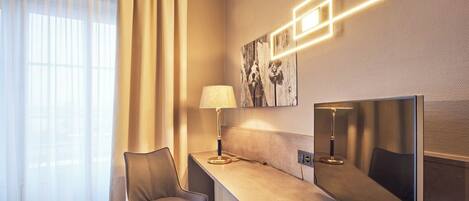 Hotelkamer met een grote flatscreen televisie, een bureau met lamp, een stoel en een raam met dunne gordijnen.
