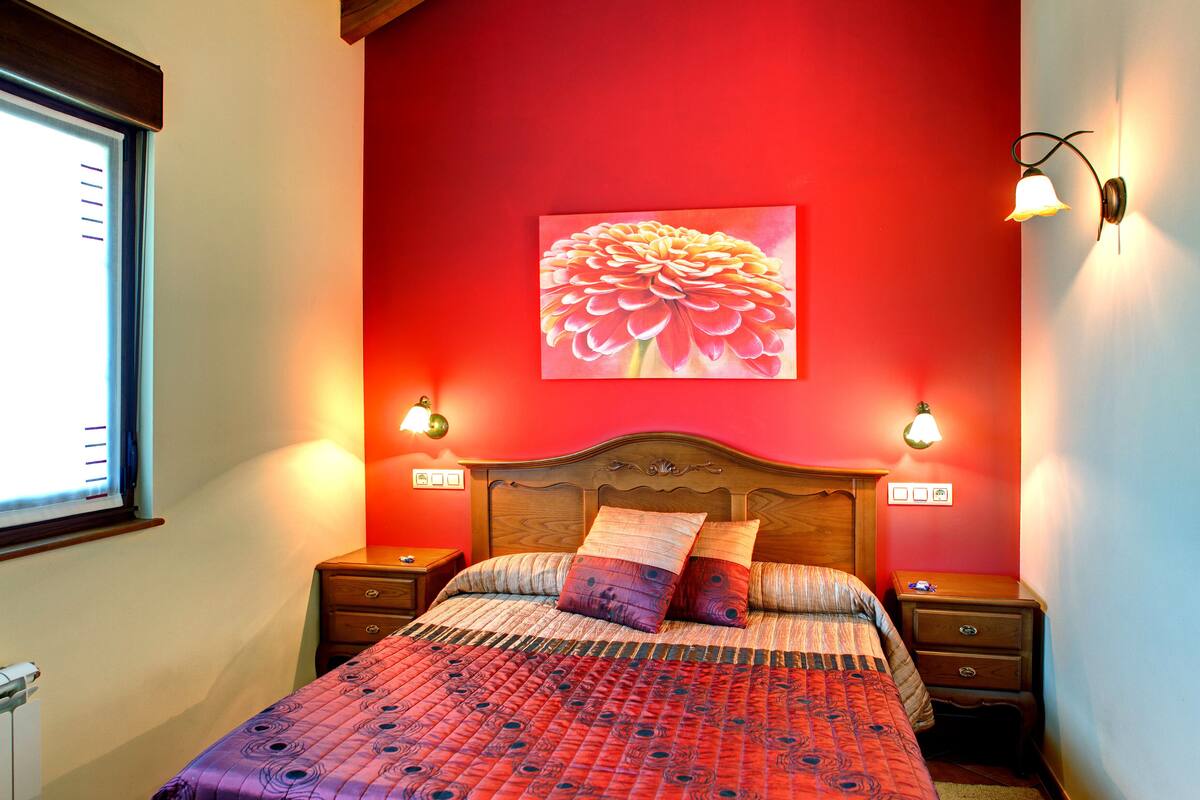 Une chambre avec un mur rouge, une tête de lit en bois, un tableau représentant des fleurs, deux lampes de chevet et une couvre-lit à motifs.