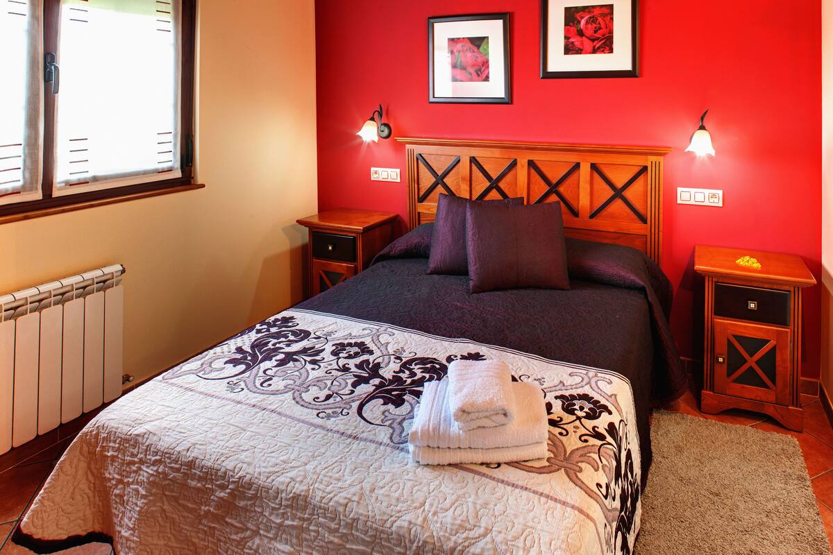 Une chambre à coucher avec un lit, deux tables de chevet, un radiateur et une fenêtre avec des rideaux.