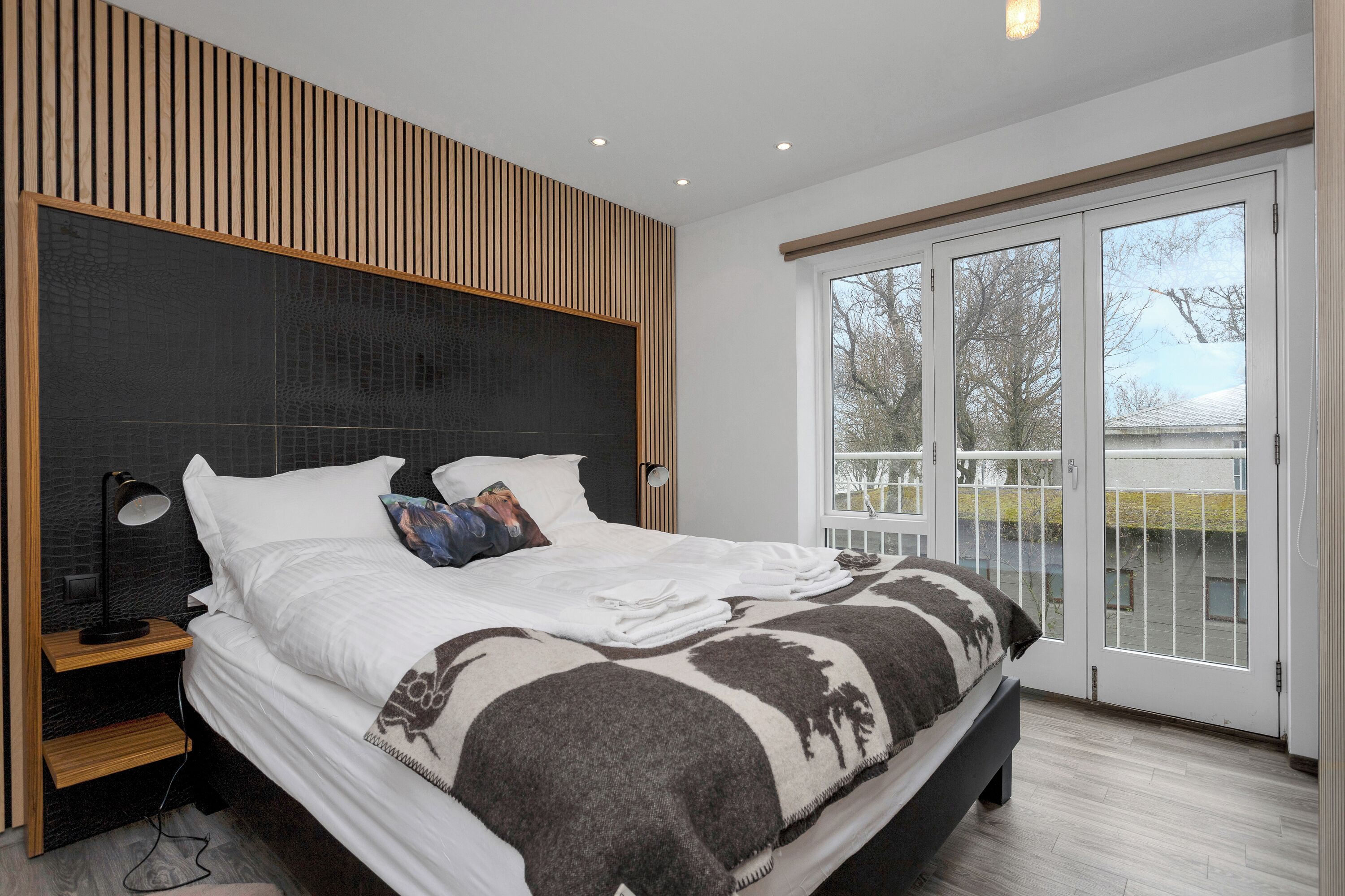 A modern bedroom with a large bed, a wooden headboard, a nightstand, and a view through a glass door to the outside.