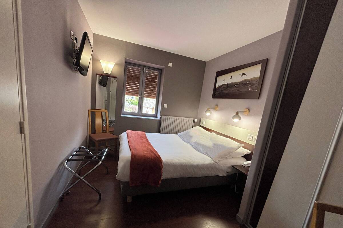 A hotel room with a bed, a chair, a desk, a window with blinds, and a painting on the wall.