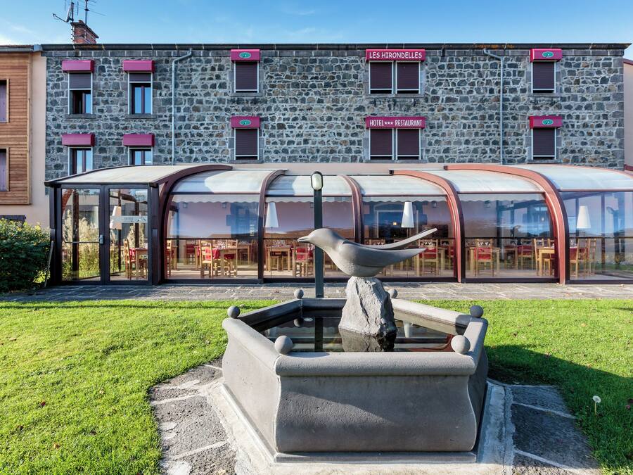 A stone building with a restaurant and hotel sign, a covered outdoor seating area, and a water feature with a bird sculpture.
