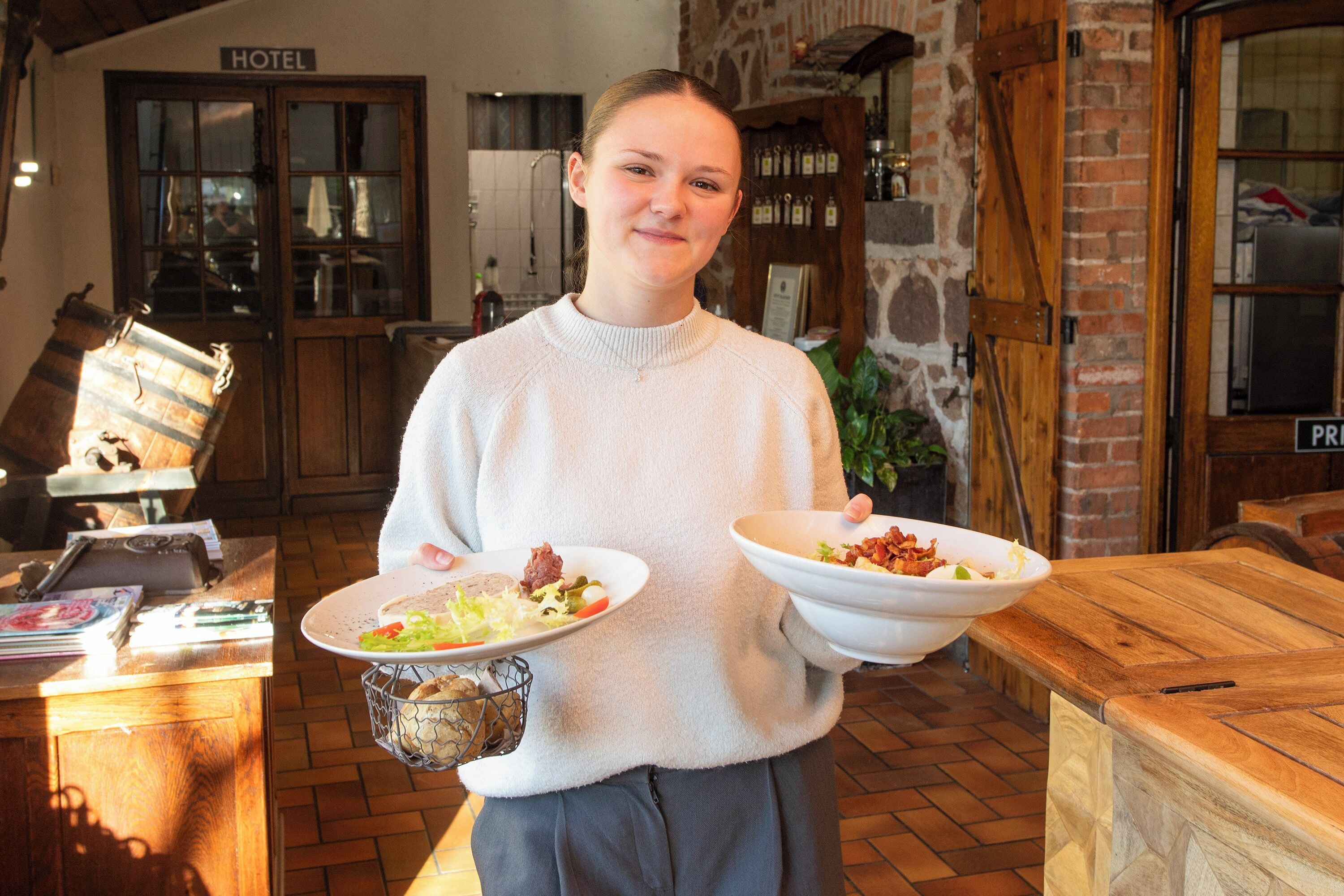 A person holding two plates of food in a room with a brick wall, wooden counter, and a sign that reads "HOTEL".