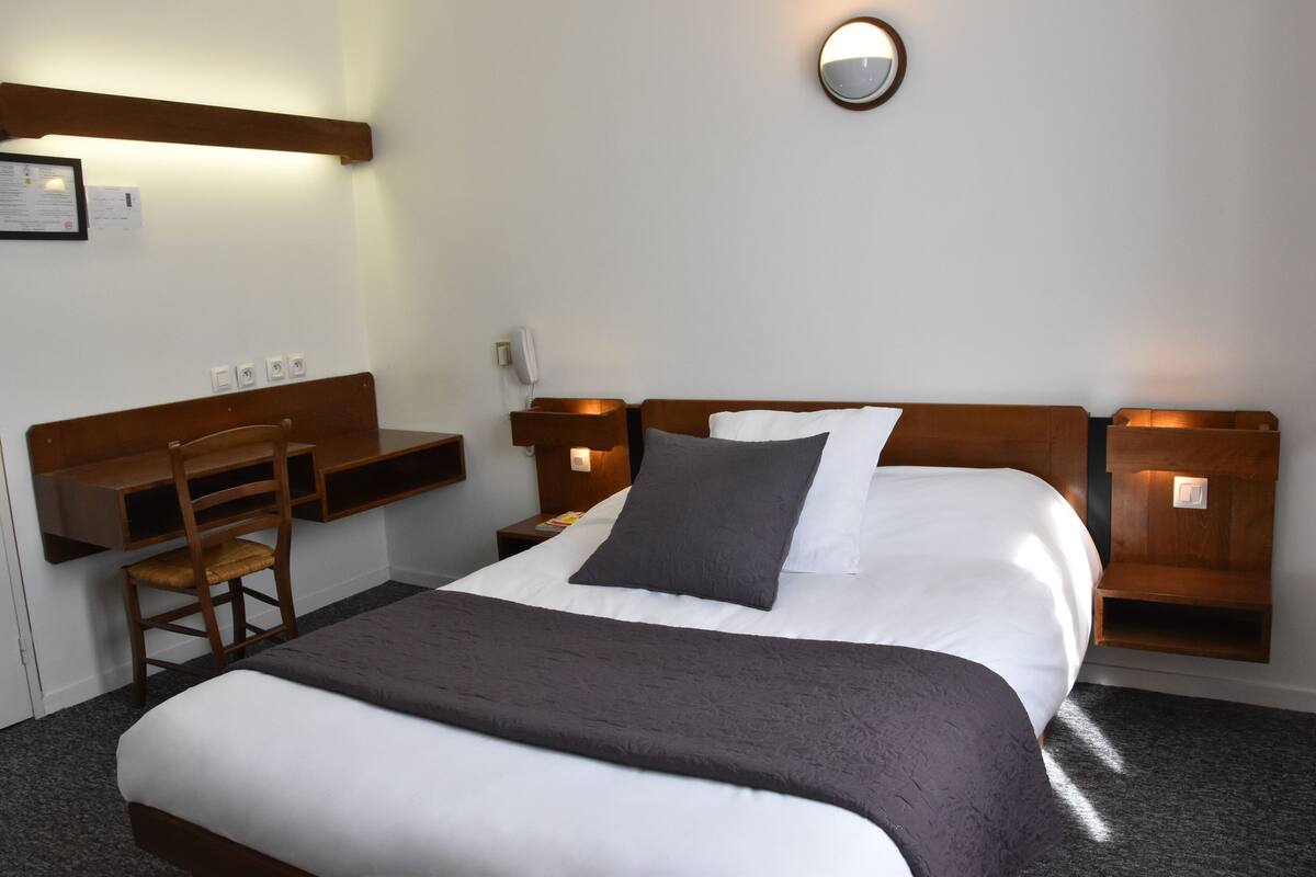 Quarto de hotel com uma cama, escrivaninha, cadeira e prateleira na parede.