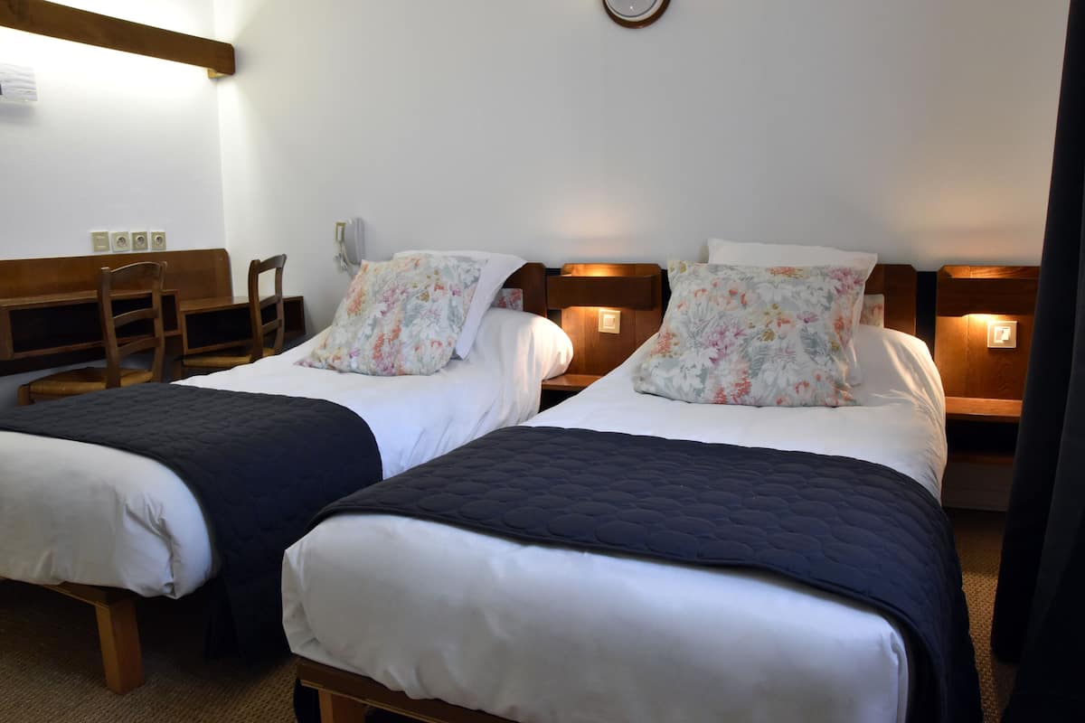 Quarto de hotel com duas camas, cada uma com cabeceira de madeira, roupa de cama branca e almofadas com padrão floral. Há mesas de cabeceira com luminárias e um banco de madeira.