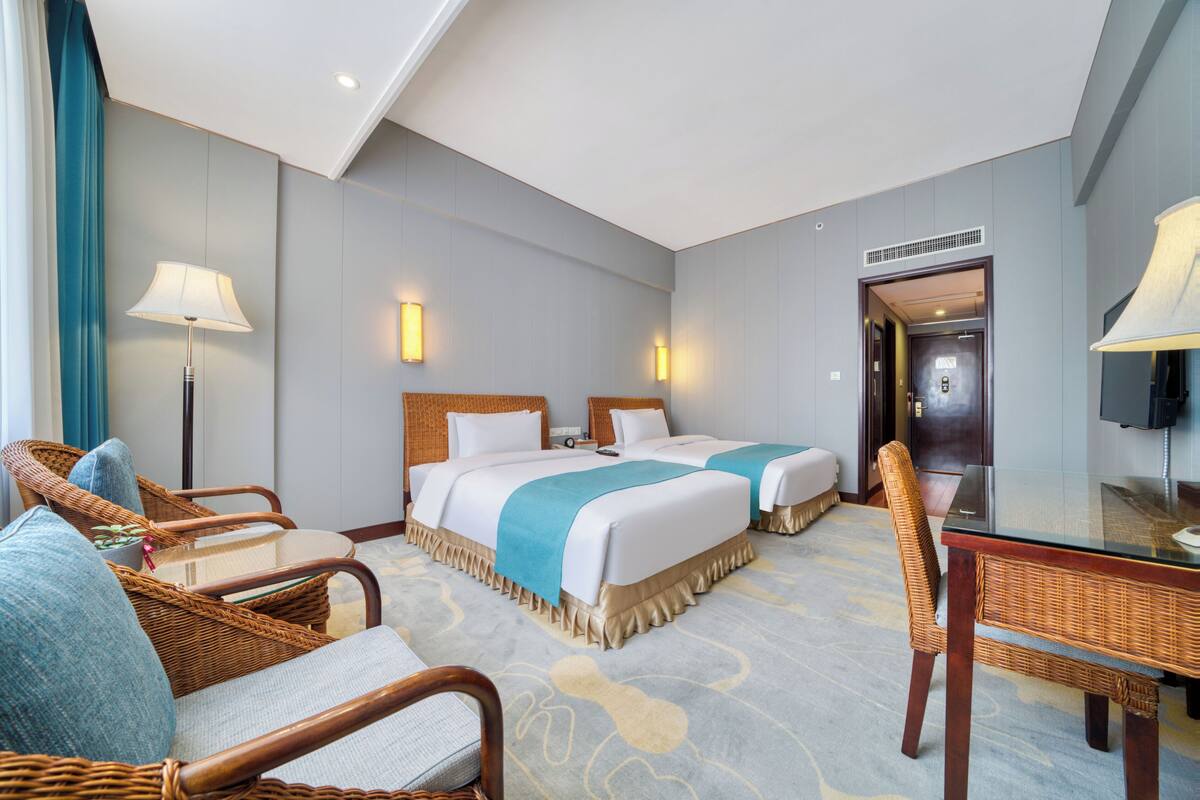 Habitación de hotel con dos camas, una zona de estar con mobiliario de mimbre, una mesa de vidrio y una televisión.