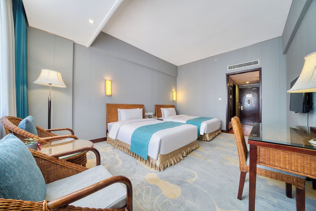 Habitación de hotel con dos camas, una zona de estar con mobiliario de mimbre, una mesa de vidrio y una televisión.