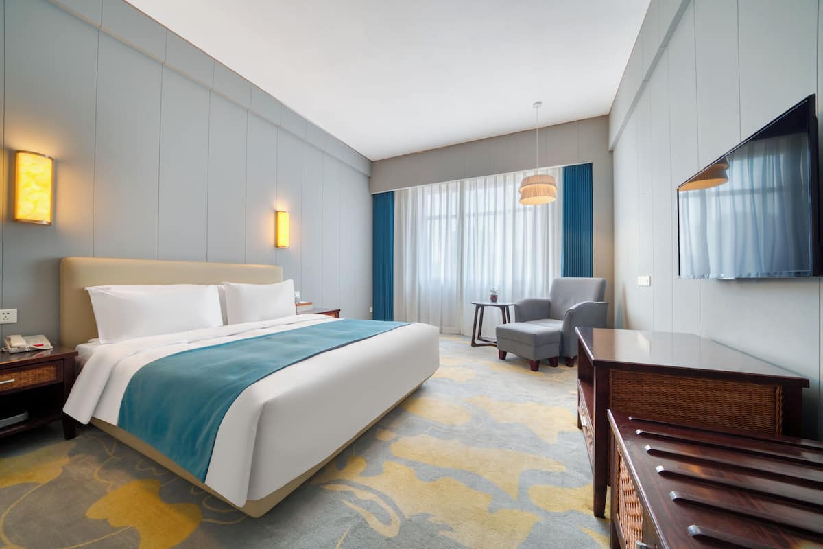 Habitación de hotel con una cama grande, un escritorio, una silla, un televisor y un ventanal con cortinas.