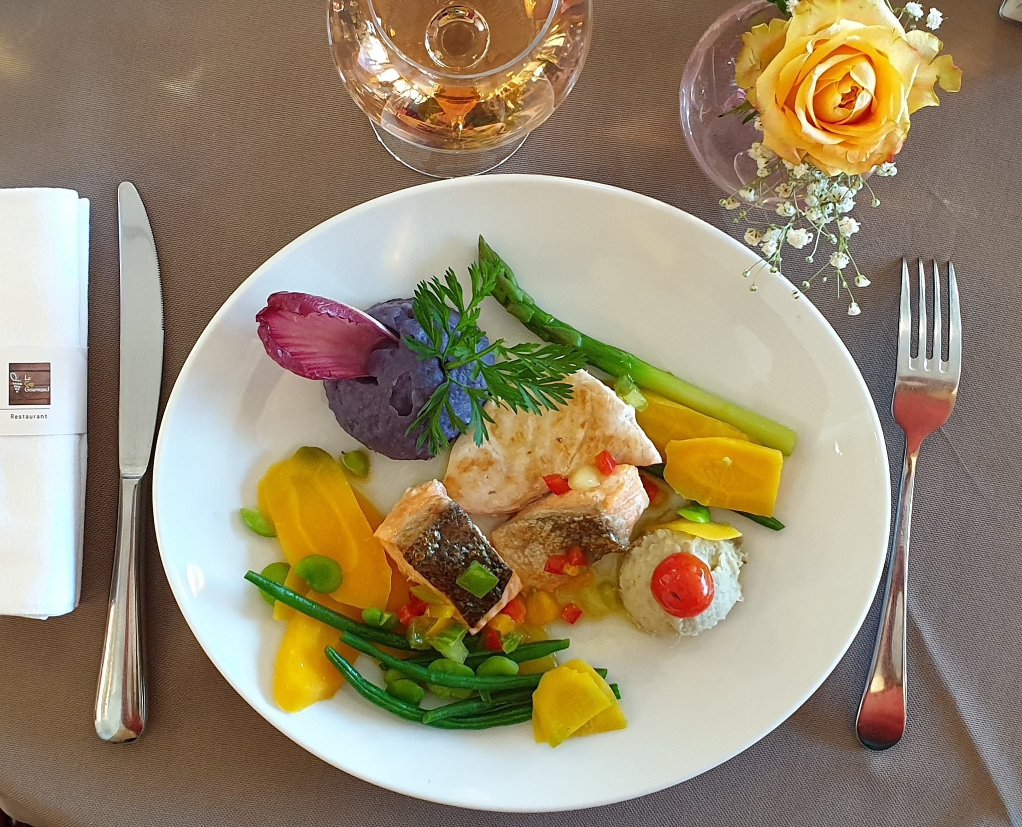 A plate of food with various vegetables and a piece of fish, accompanied by a glass of wine and a vase with a yellow rose.