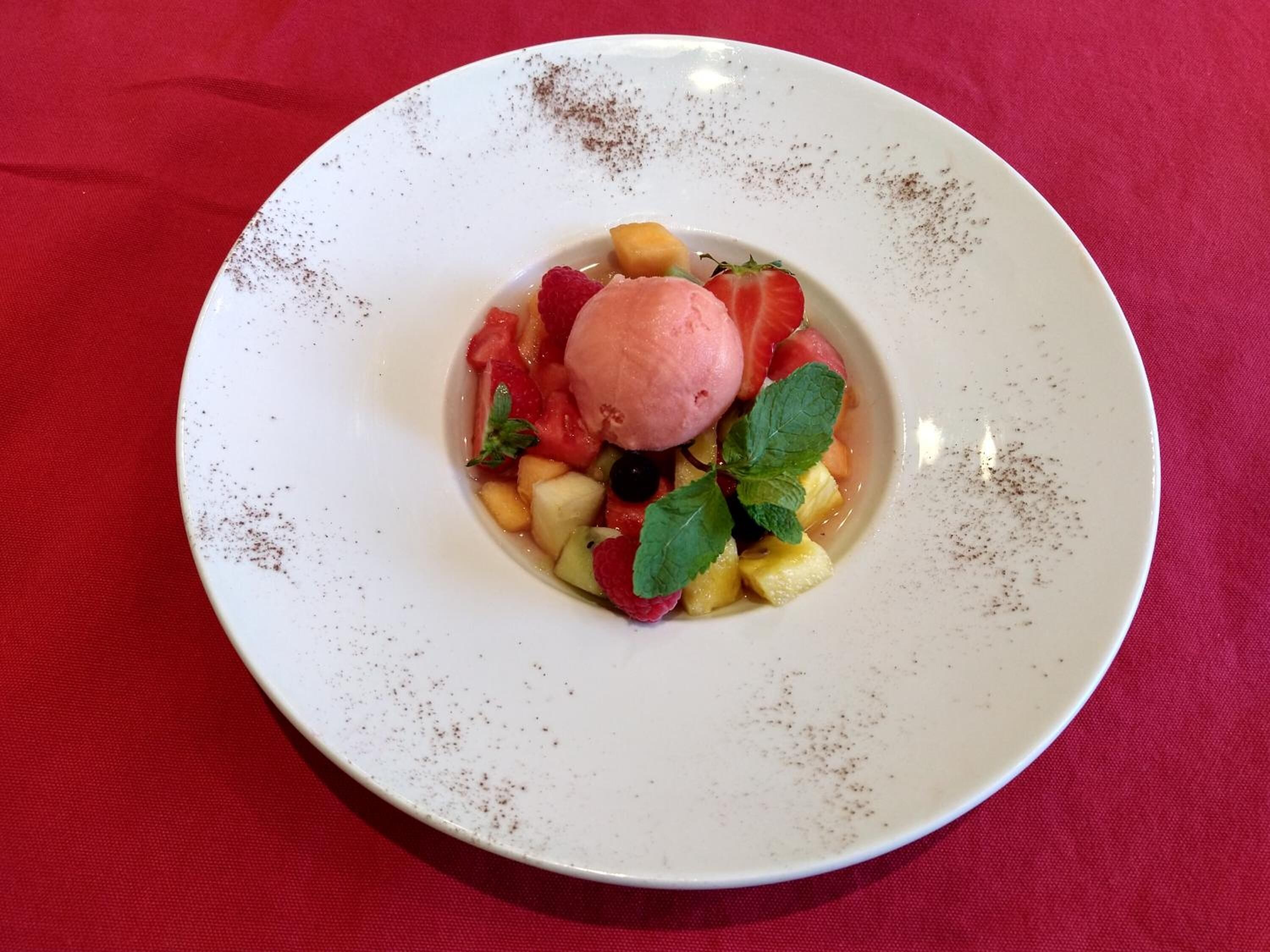 Un dessert servi sur une assiette blanche, agrémenté de fruits et de menthe, le tout posé sur une nappe rouge.