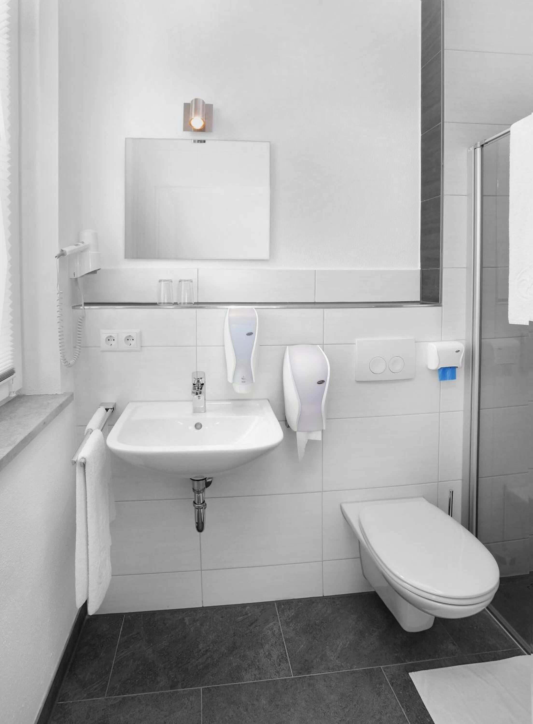 A modern bathroom with a white sink, mirror, and toilet. There is a towel rack, a soap dispenser, and a shower area.