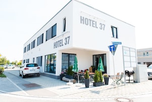 A modern hotel building with the sign "HOTEL 37" and "Bernsteinstraße" visible. There are outdoor seating areas with umbrellas and potted plants.