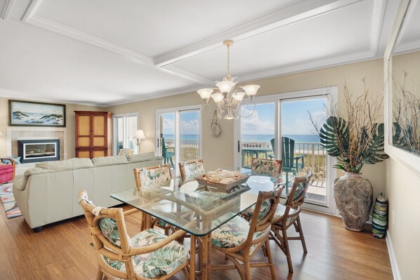A spacious living room with a glass dining table, wicker chairs, a sofa, and a view of the ocean through large windows.