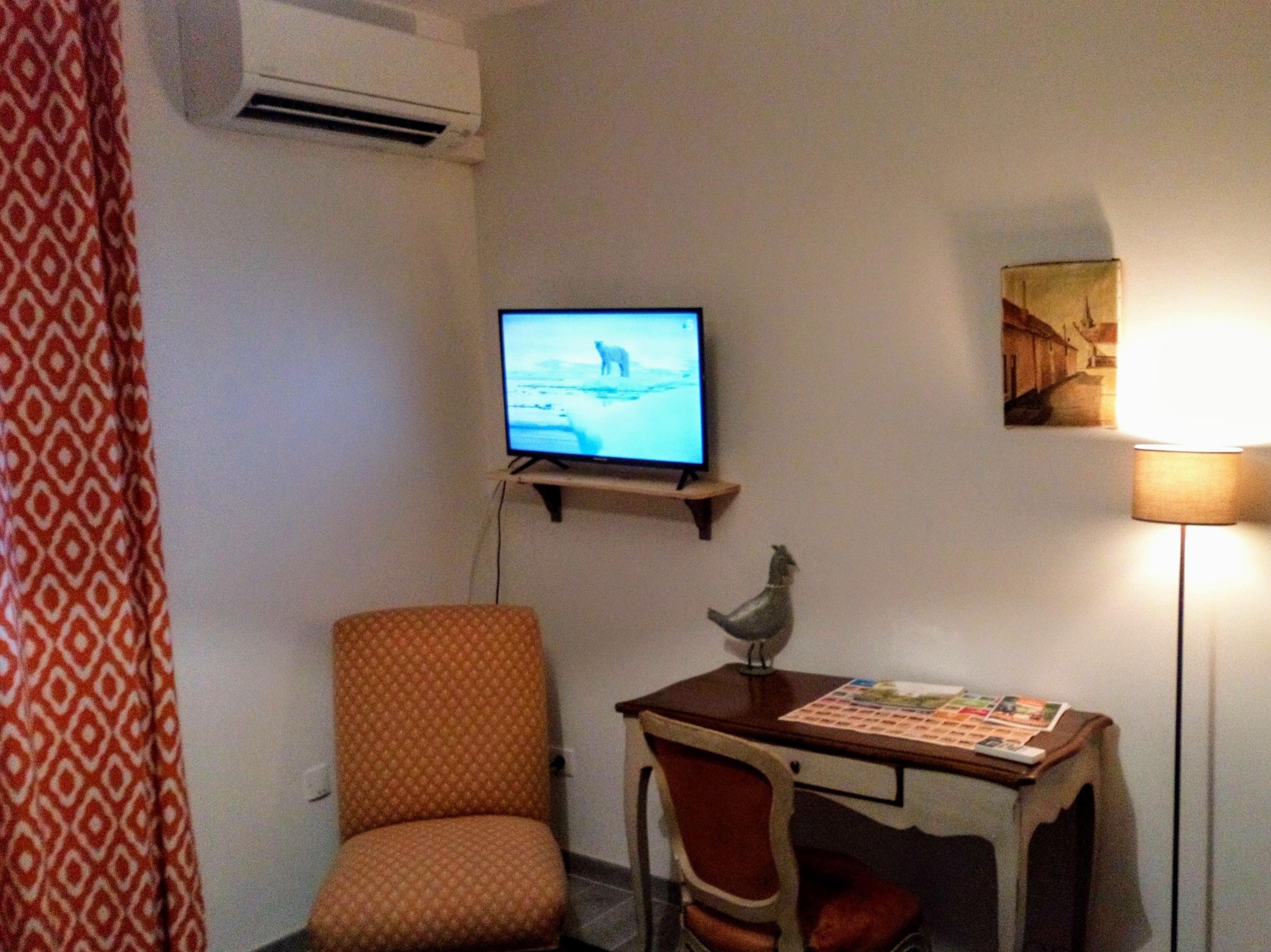 Una habitación con una televisión instalada en la pared, una silla, una mesita con una lámpara y un aire acondicionado también fijado a la pared.