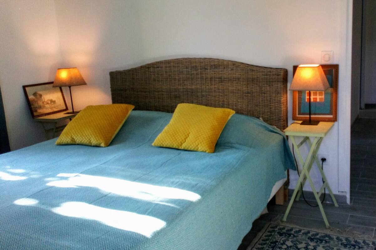 Uma cama com colcha azul e duas almofadas amarelas, com duas mesas de cabeceira de cada lado, cada uma com uma luminária.