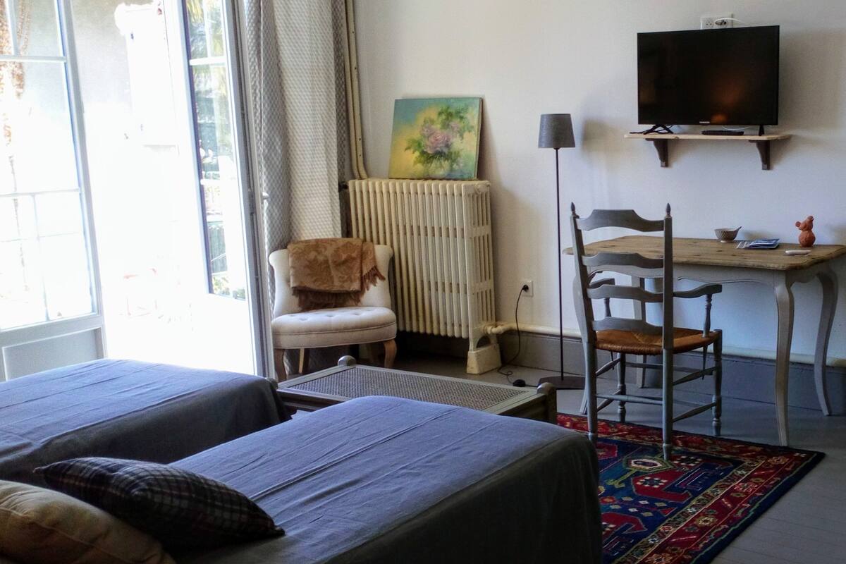 Quarto com duas camas, uma escrivaninha, uma cadeira, um radiador, uma televisão e uma janela com cortinas.