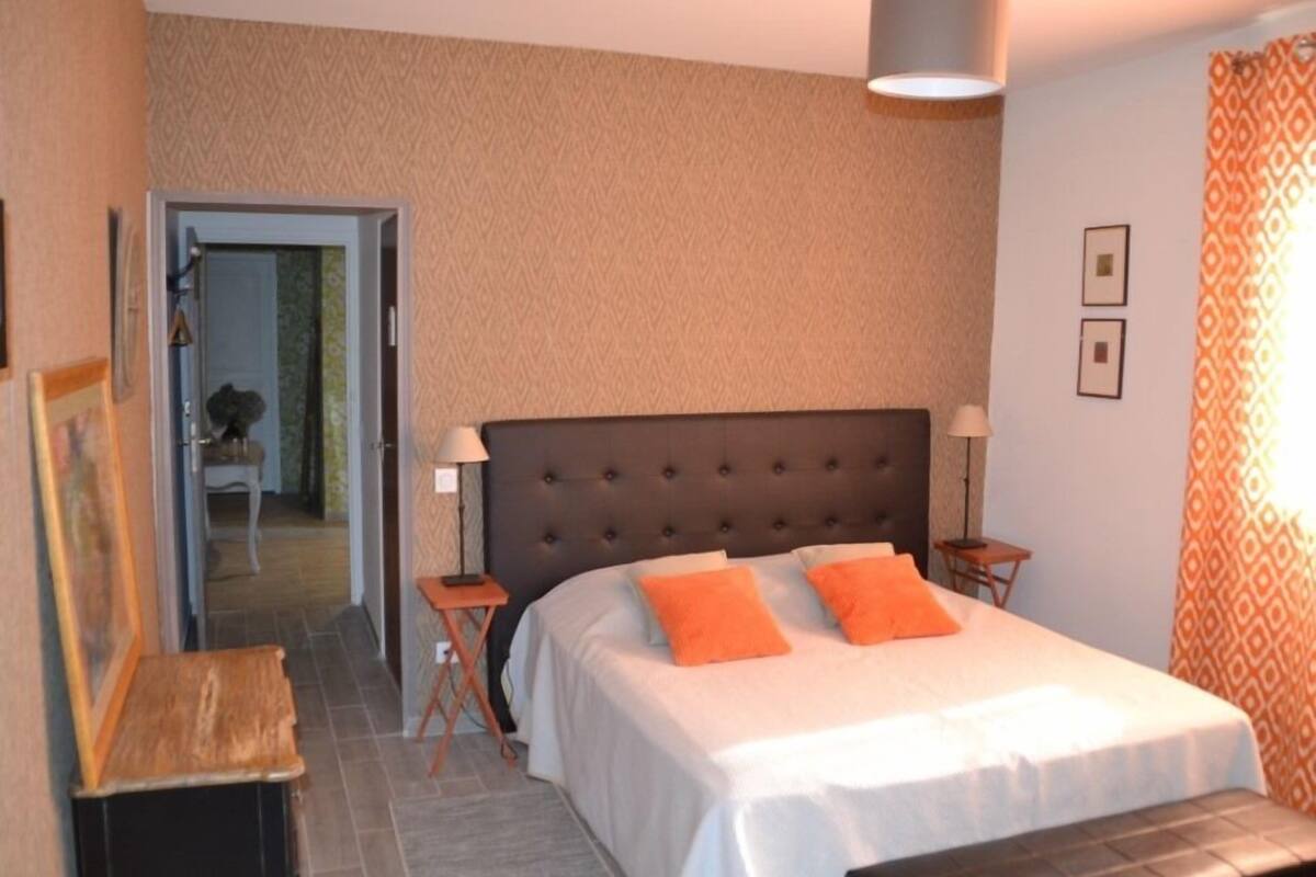 Quarto com uma cama grande, duas mesas de cabeceira, um criado-mudo, um banco e uma porta que dá para outro cômodo.