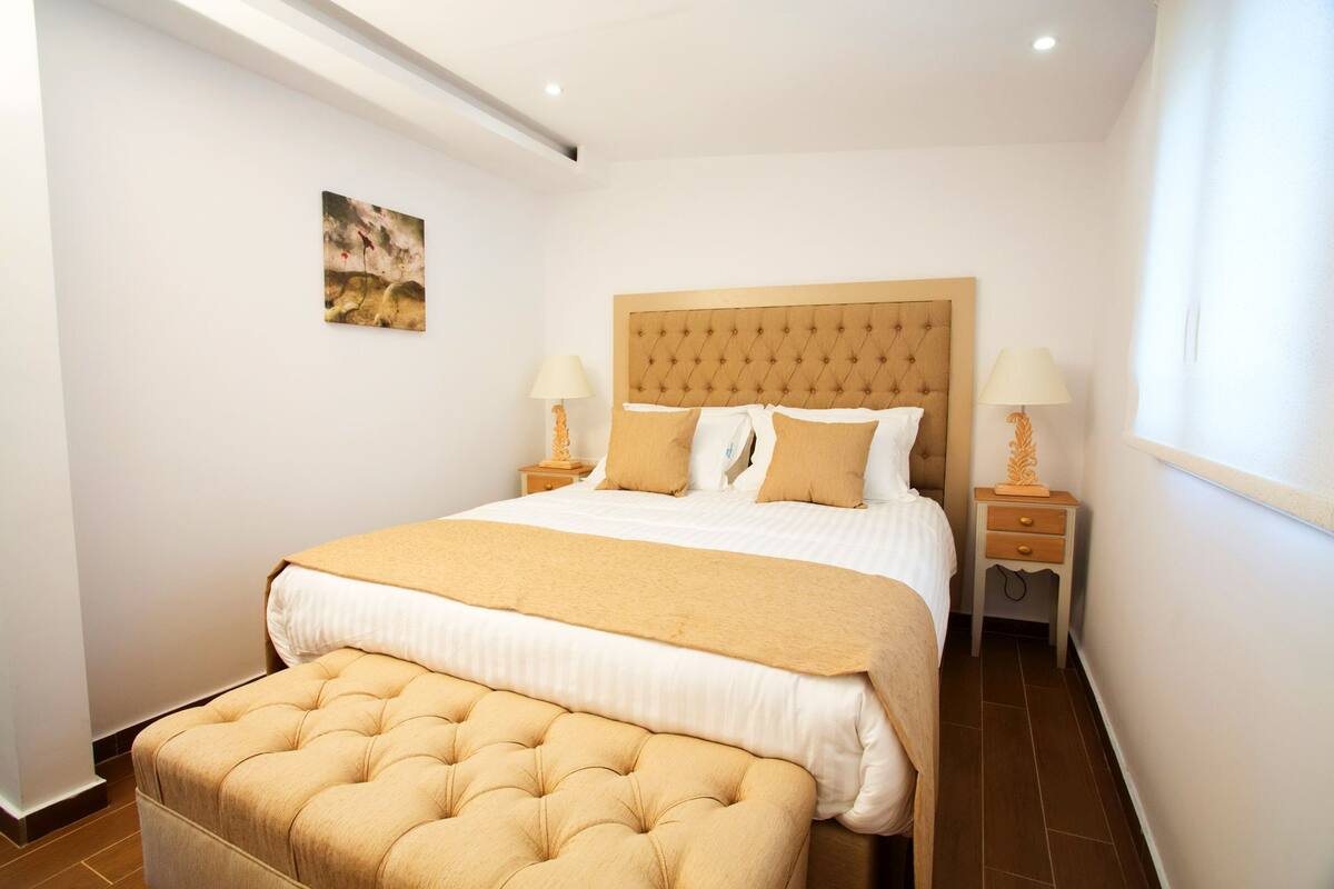 Un dormitorio con una cama grande, dos mesitas de noche con lámparas, una obra de arte pequeña en la pared y una ventana con cortinas.