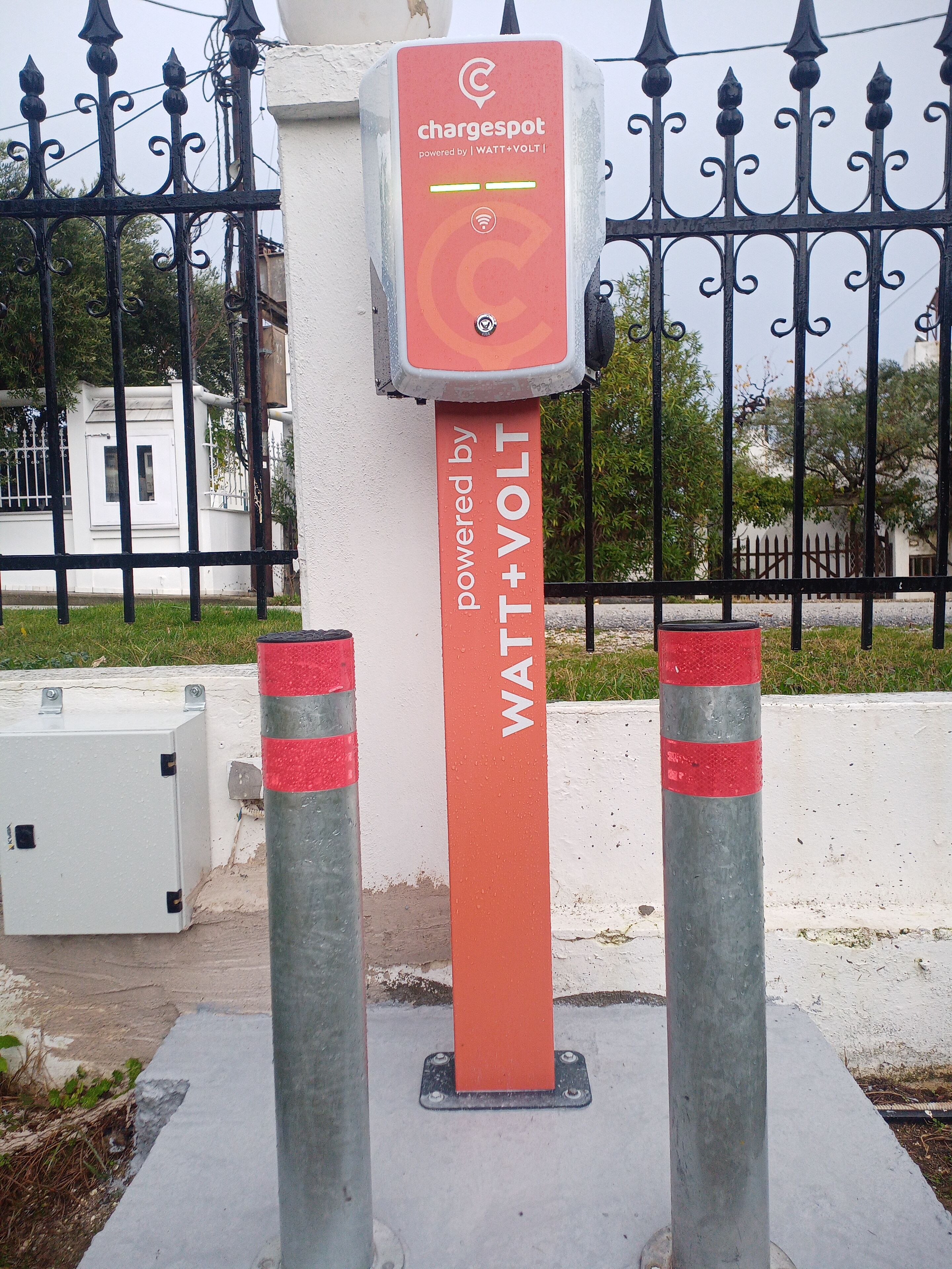 Una estación de carga pública con el rótulo 'chargespot powered by WATT+VOLT' y una cerca metálica al fondo.