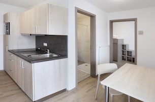 Cozinha moderna com armários de madeira clara, bancada preta e pia de aço inoxidável. Há uma cadeira branca e uma mesa na área de jantar adjacente.
