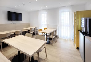 Quarto de hotel moderno com uma grande TV de tela plana, várias áreas de estar com mesas e cadeiras, e uma kitchenette com micro-ondas.