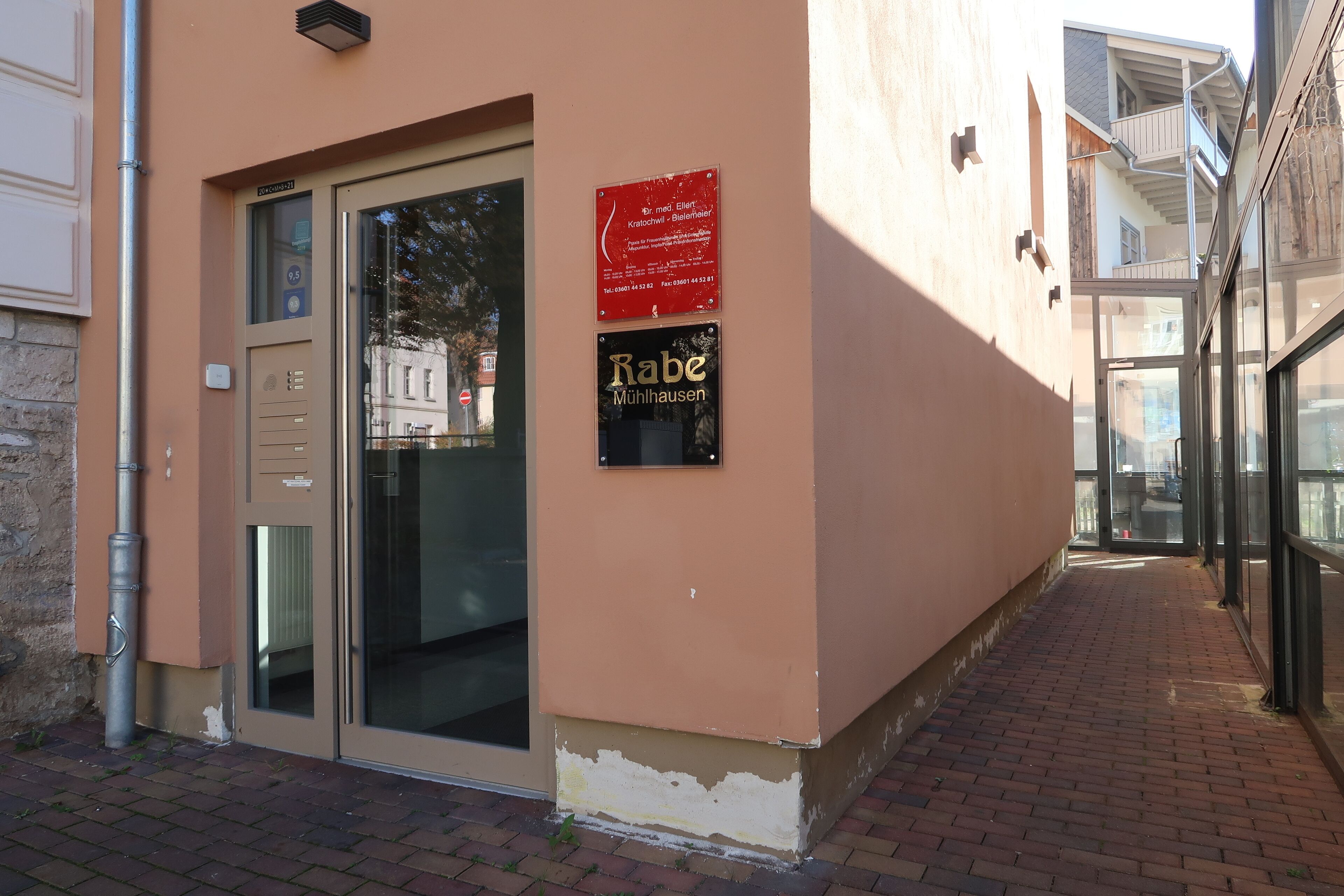 Un edificio con puerta de entrada de vidrio, un letrero con información de contacto y una placa que indica el nombre 'Rabe Mühlausen'.