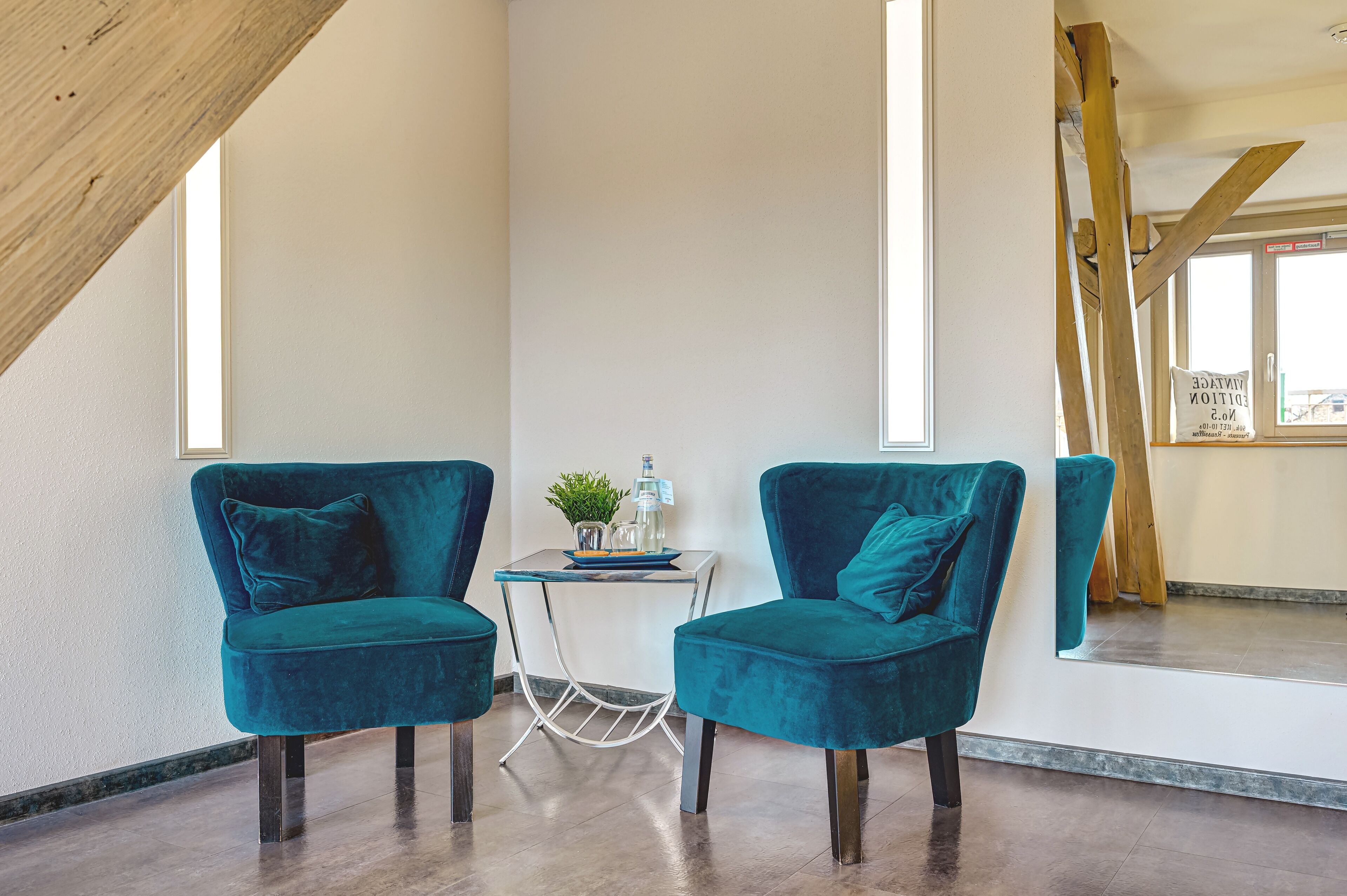 Una habitación con dos sillones de terciopelo color turquesa, una mesita con una planta y una ventana con estructura de vigas de madera.