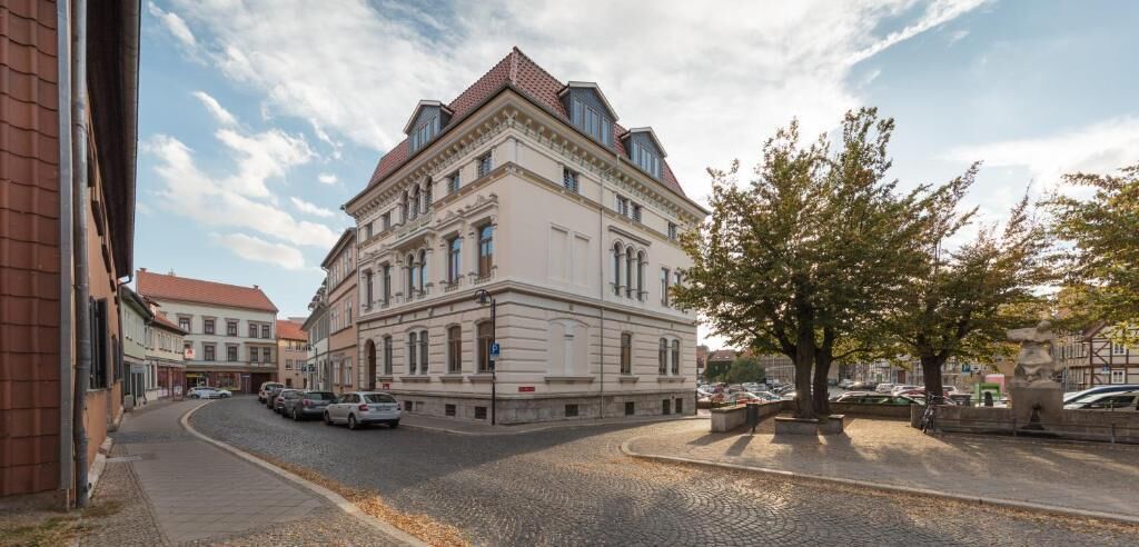 Ein historisches Gebäude mit klassischer Architektur, das einen markanten Hauseingang an der Ecke und einen großen Baum davor zeigt.