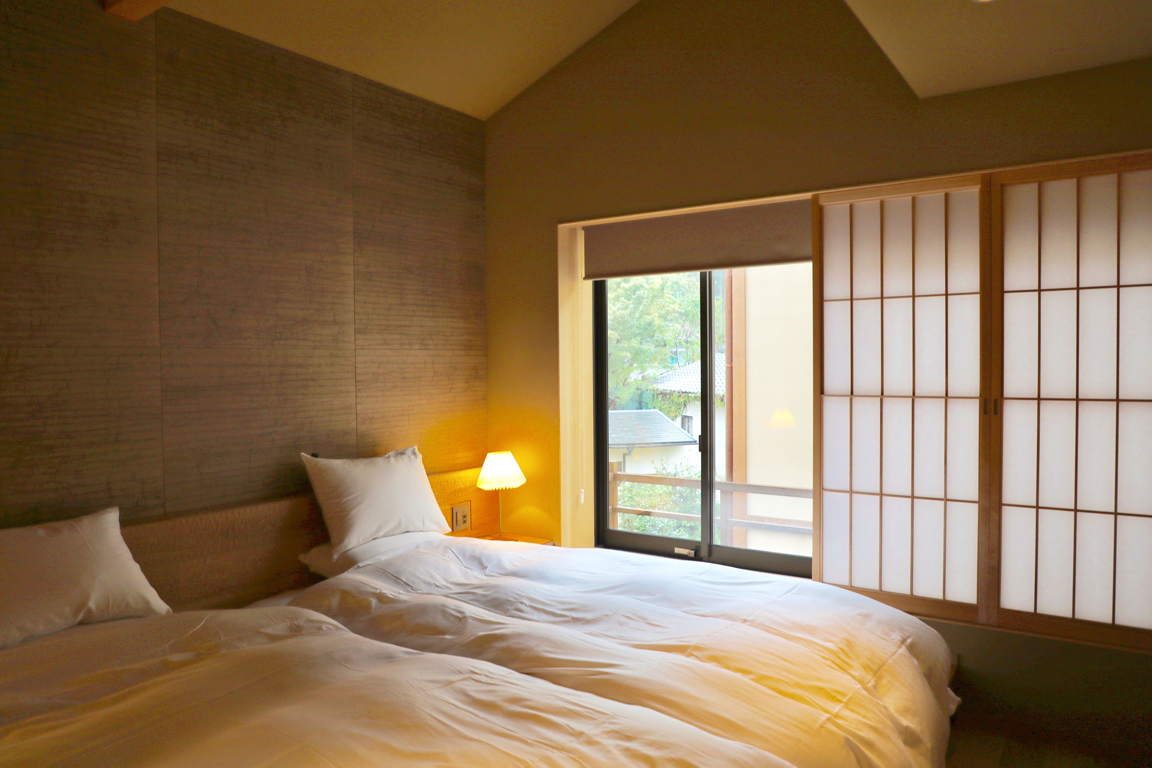 A hotel room with a large bed, a nightstand with a lamp, a sliding shoji screen, and a view of the outdoors.
