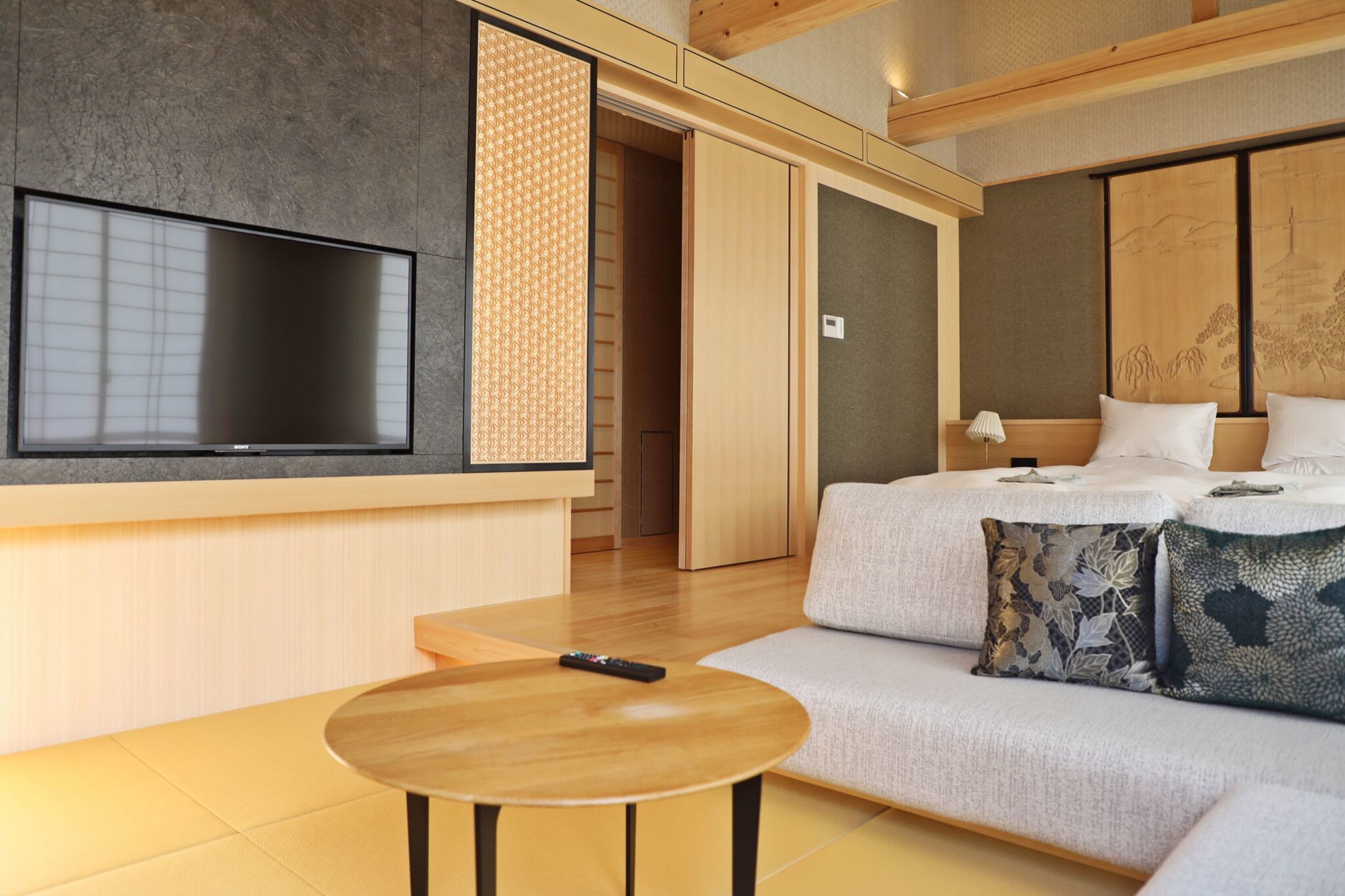 A modern hotel room with a large flat-screen TV, a sofa with decorative pillows, a wooden table, and a sliding door leading to a bathroom.