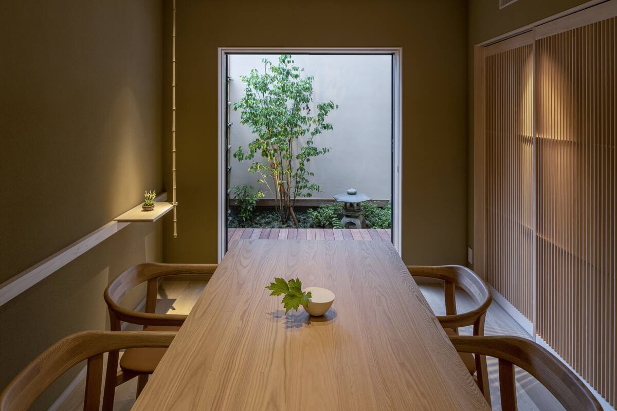 A dining area with a long wooden table, chairs, a small plant, and a view of a tree through a doorway.