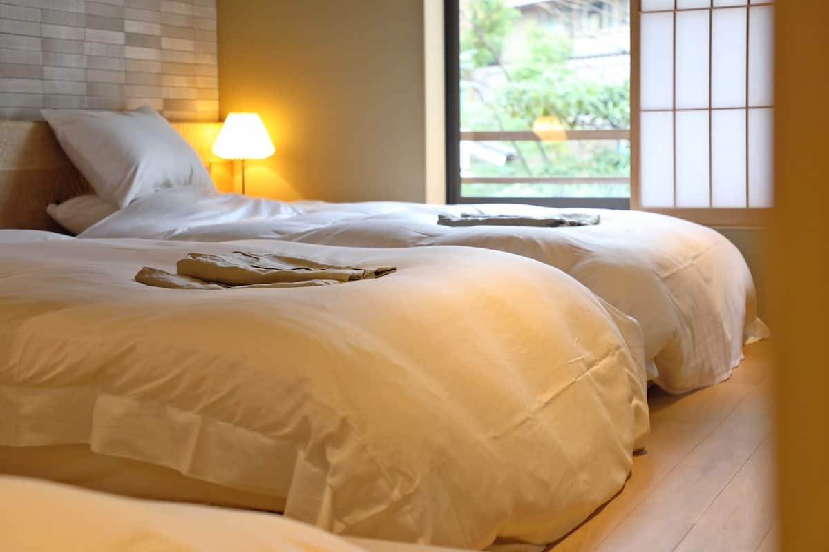 A double bed with white linens, a bedside lamp, and a view of greenery through a window.