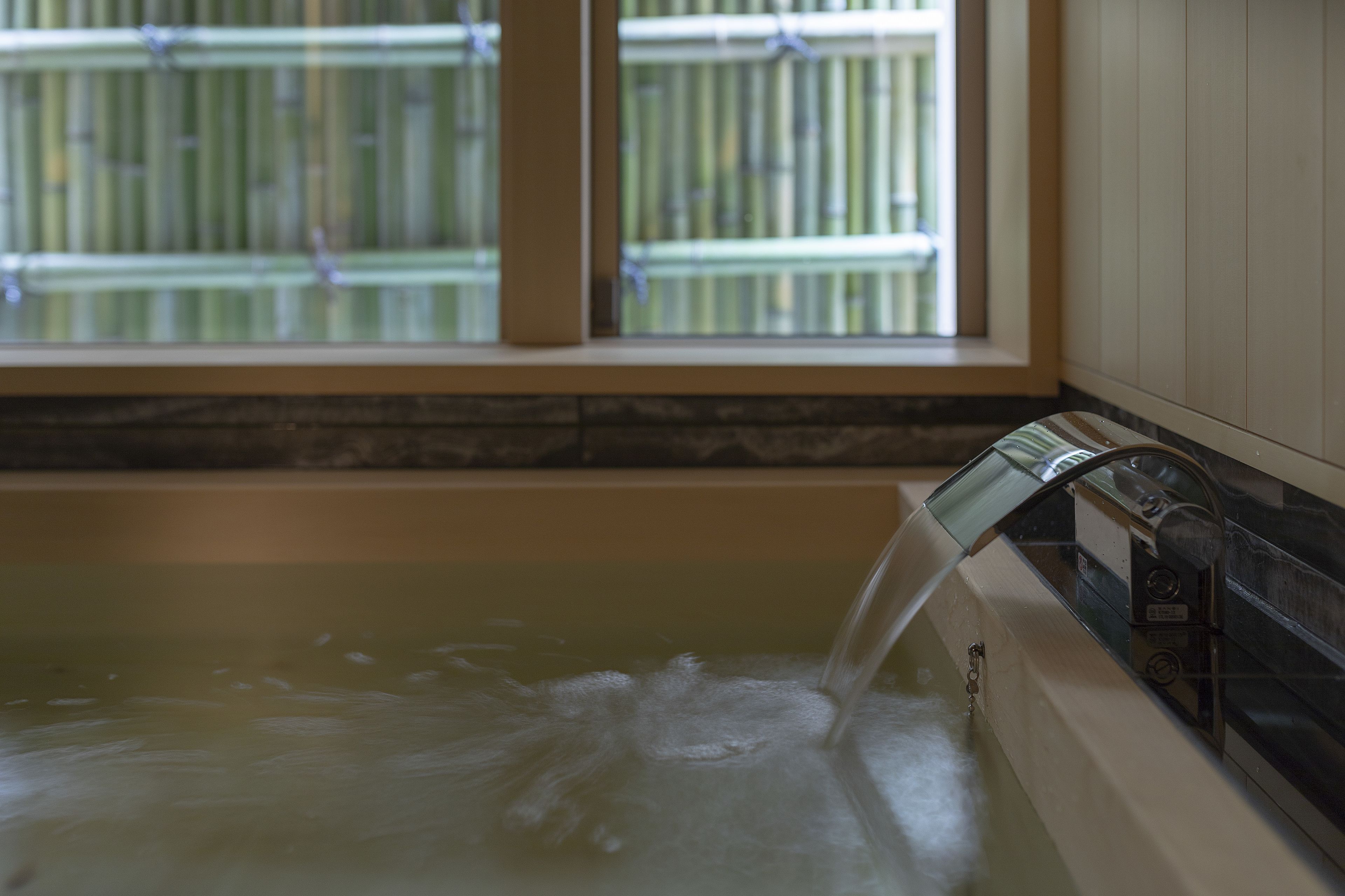 A bathtub with running water, a window with a view of a bamboo fence, and a stone countertop.