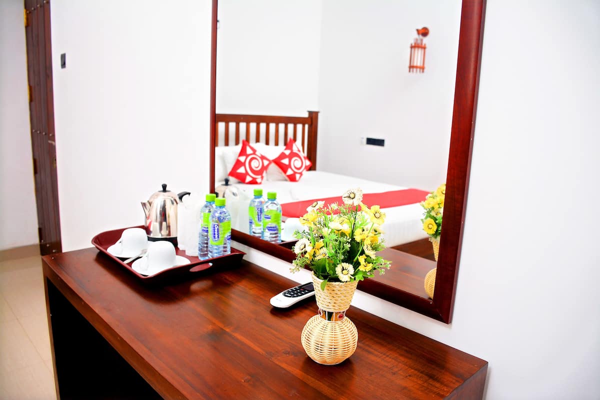 A hotel room with a wooden desk, a vase of flowers, a tray with a kettle and cups, and a bed with red and white pillows.