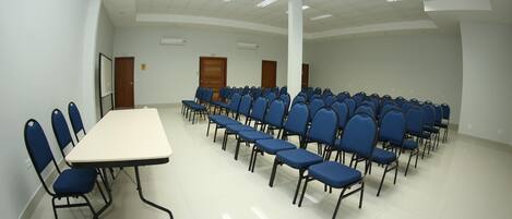 A conference room with a long table and rows of blue chairs, white walls, and a tiled floor.