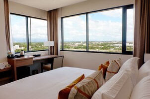 Habitación de hotel con una cama grande, un escritorio y vistas a un paisaje urbano a través de amplios ventanales.