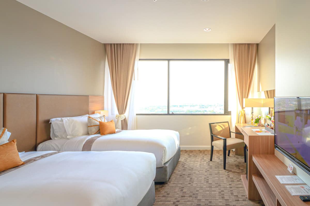 Quarto de hotel com duas camas, uma escrivaninha, uma cadeira, uma televisão e uma janela grande com cortinas.