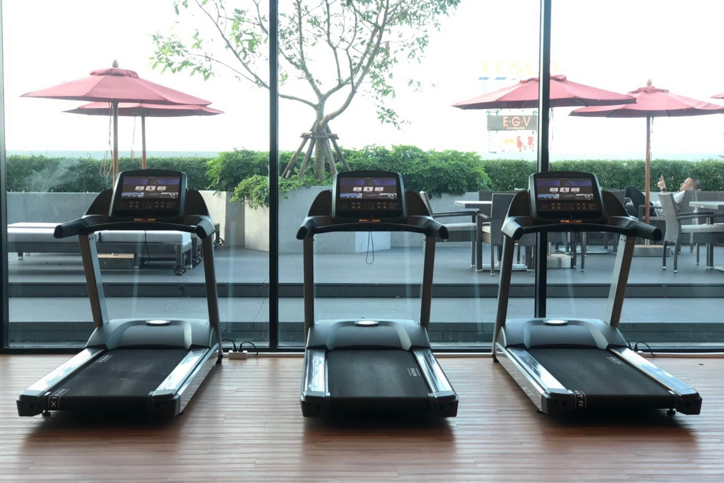 Three treadmills in a room with large windows overlooking a patio area with umbrellas and outdoor seating.