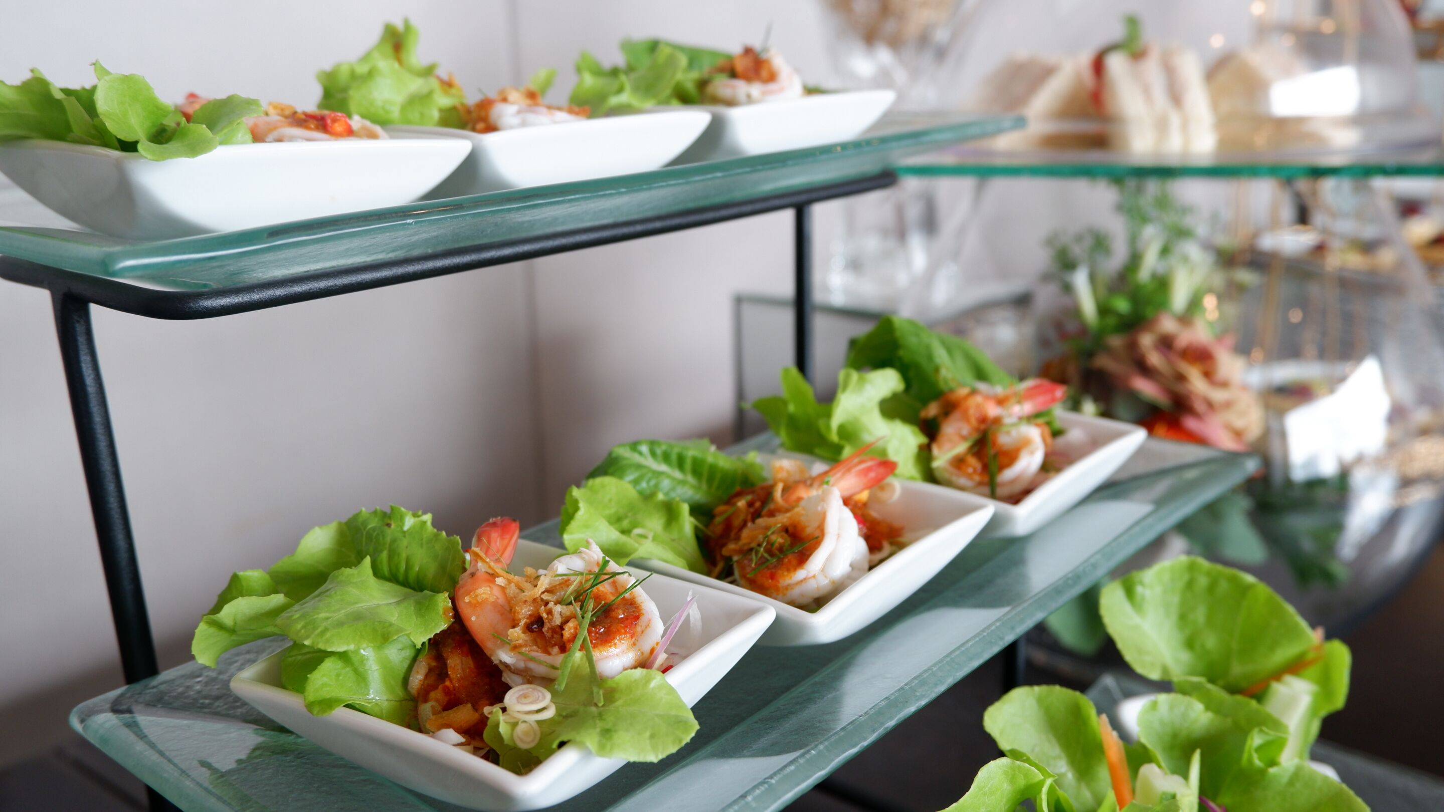 A buffet table with shrimp appetizers on white square plates, garnished with lettuce and herbs.