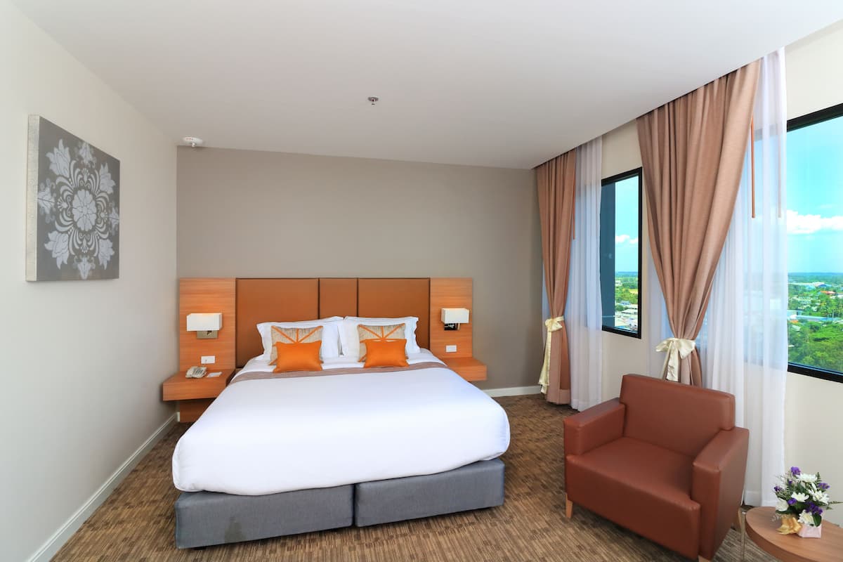 Quarto de hotel com uma cama grande, cabeceira de madeira, duas mesas de cabeceira com luminárias, uma poltrona marrom e uma janela com cortinas.