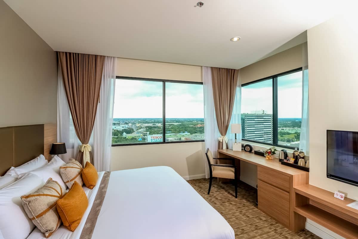 Quarto de hotel com cama, escrivaninha, cadeira, TV e janelas amplas com vista para a cidade.