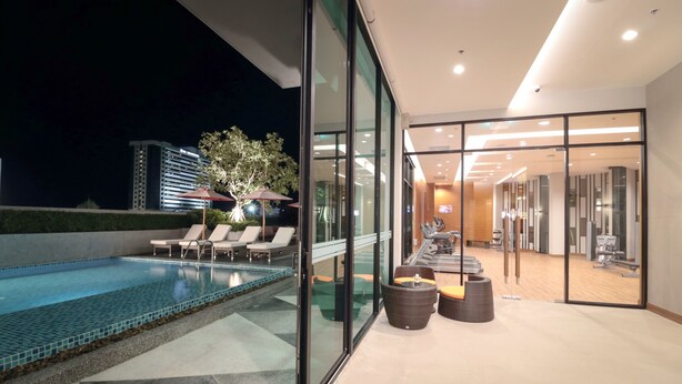 A modern hotel pool area with a glass-enclosed spa room, lounge chairs, and exercise equipment. - Grand Fortune Hotel Nakhon Si Thammarat (Nakhon Si Thammarat)